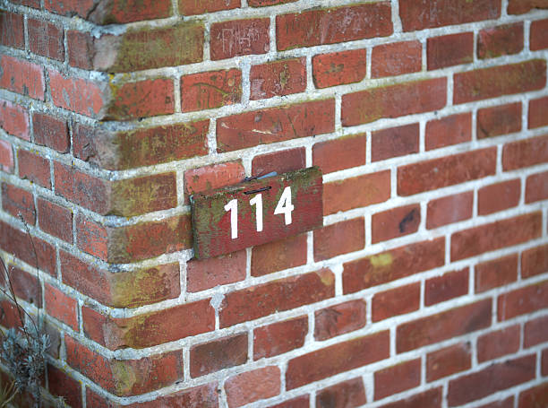 A brick wall corner with a wooden number plate marked 114, showcasing the rustic charm of Danish architecture. Weathered bricks are arranged with visible patterns, emanating historical and rural appeal.
