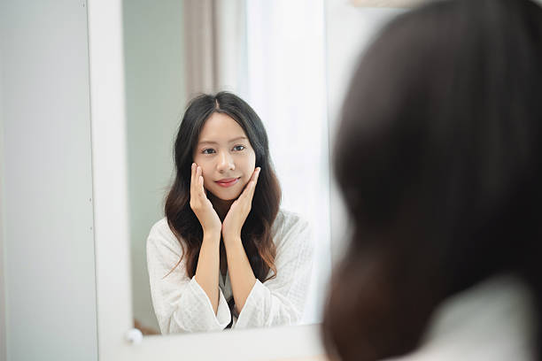 istock Young woman in white robe admiring her reflection in a mirror while touching her face, showcasing self-care and beauty routine in bright room 2229178516
