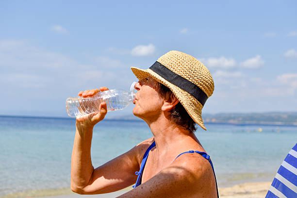Beautiful young woman in swimsuit drinks clean water from bottle, against blue teal sea and sky in sunny summer day on beach. Health care, healthy active lifestyle of modern people concept.
