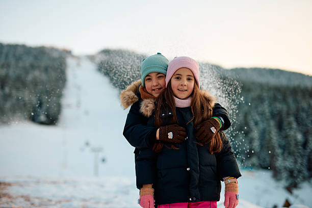 Two children are hugging and smiling at the camera in a snowy mountain landscape, wearing winter clothes and gloves, with snow falling around them