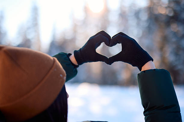 Woman creating a heart shape with her gloved hands in a snowy forest, basking in the warm glow of sunset. Nature's beauty enhances the romantic winter landscape