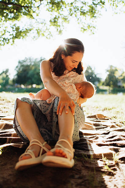 A serene moment of a mother nursing her infant while seated on a blanket under dappled sunlight in a lush park, capturing the beauty of maternal care and nature
