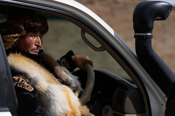 Environmental portrait of a man wearing a traditional fur hat and embroidered winter coat, seated at the wheel of a white off-road vehicle in Mongolia. Natural sunlight highlights textures, blending tradition with modern travel. No brands visible.