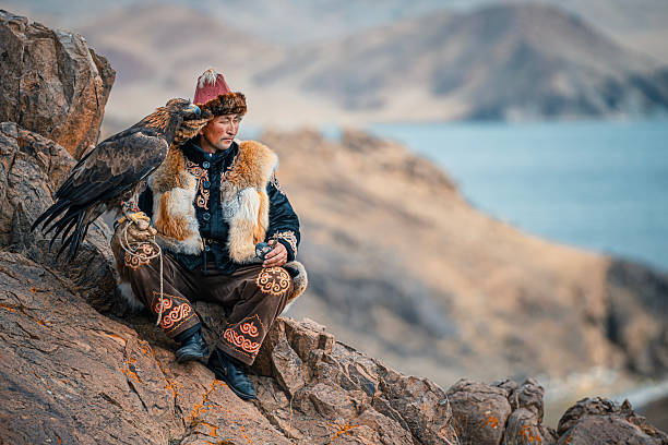 Mongolia – Kazakh eagle hunter wearing traditional fur-lined clothing, sitting on a rocky cliff with a trained golden eagle perched on his arm, overlooking a mountainous landscape near a lake. Cultural heritage, traditional hunting, and nomadic lifestyle concept.