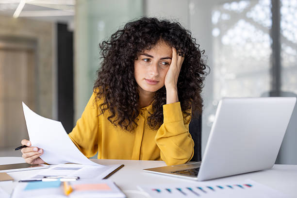 istock A focused woman in a yellow shirt reviews documents, appearing tired while working on her laptop. 2229195522
