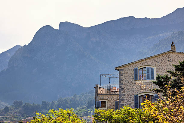 Train station on the railroad to Sóller from Palma, featuring a stone building with green shutters and a few potted plants. The area is quiet with minimal foot traffic, and a mountain landscape is visible in the background.