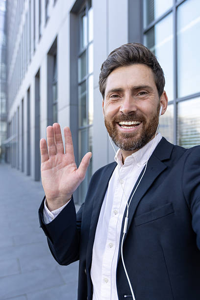 istock A happy businessman waves hello while wearing earbuds in front of a modern building. 2229206603