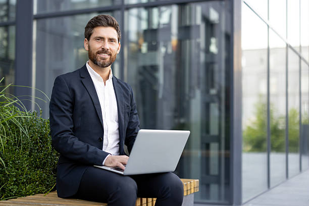 istock A well-dressed man smiles while using a laptop outside a modern office building. 2229206670