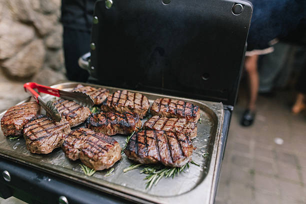 Juicy grilled steaks with seared lines arranged on a barbecue. Stainless steel tongs and green herbs enhance the culinary display. Vibrant outdoor ambiance during social gathering, ideal for summer grilling enthusiasts