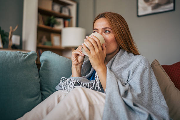 Cozy portrait of a sick mid adult woman wrapped in a blanket, enjoying a warm drink on the sofa in her living room, finding comfort and relief