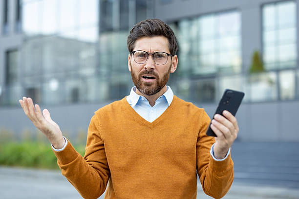 istock A businessman looks perplexed while holding a smartphone, outside near a building. 2229212361