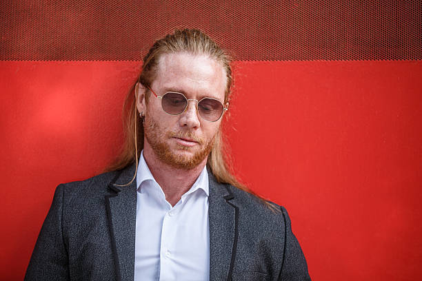 A Redhead adult man with long hair, wearing a business suit and sunglasses, leans against a red wall, appearing thoughtful. Shot in outdoor urban area during the daytime.
