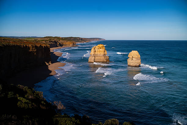 A silhouetted couple holds hands while standing on the beach at sunrise, with the towering limestone stacks of the Twelve Apostles in the background along Victoria’s iconic Great Ocean Road.  The Twelve Apostles viewed from the beach at sunrise glow with golden hues as morning light reflects off the sandstone. Access to the beach beneath the Twelve Apostles involves a steep descent via Gibson Steps and is best attempted at low tide. Sunrise is one of the most photogenic times, offering both solitude and spectacular lighting. Visitors are advised to arrive early and bring a flashlight if descending before first light.