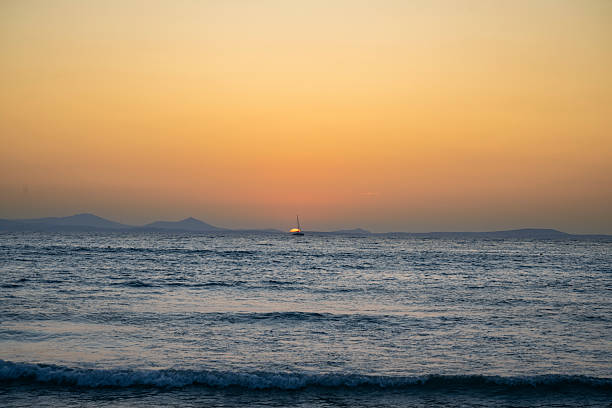 A sailboat drifts across the Aegean Sea as the sun sets behind distant islands, casting warm golden and orange hues over the tranquil waters off Naxos, Greece.