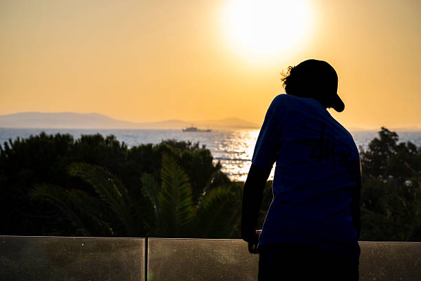 Silhouette of a young man leaning on a glass railing, gazing at the Aegean Sea during sunset. Warm light and distant ship create a peaceful summer mood.
