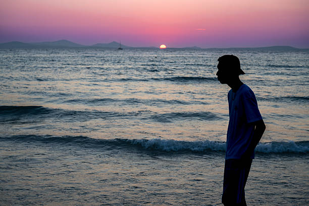 Silhouette of a young man walking along the shore at sunset, with pink and purple skies reflecting over the calm Aegean Sea on Naxos, Greece.