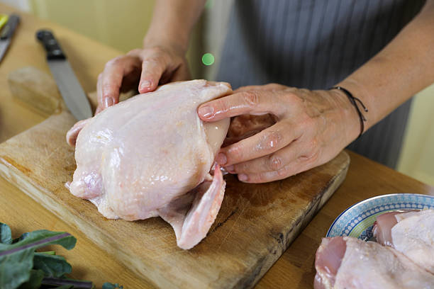Woman in a Red T-shirt and Gray Apron Slices a Raw Chicken in the Kitchen. The Scene Captures the Precision and Care of a Woman Preparing a Meal, Surrounded by Kitchen Tools and Ingredients. The Atmosphere Emphasizes the Process of Cooking, Showcasing Her Focus and Culinary Skills.
