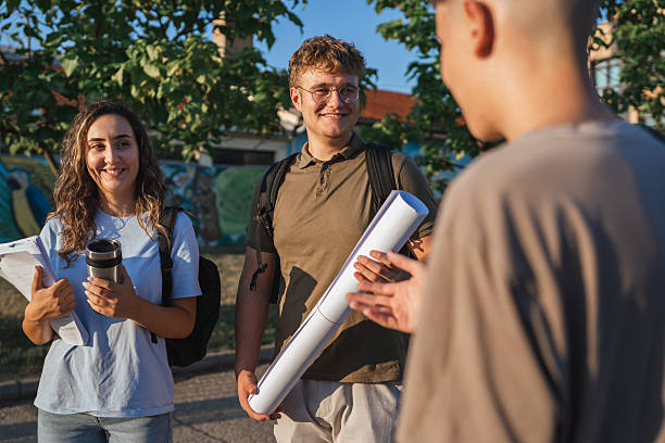 Three architecture students carrying blueprints and documents are discussing a project outdoors, engaging in a collaborative exchange of ideas