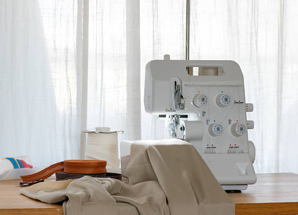 Unbranded serger overlock sewing machine operating in a home sewing studio. Female hands guide fabric under the presser foot while large thread cones feed the machine. Clean, neutral light and close-up details illustrate stitching, edge finishing and garment production. Perfect for tutorials, courses, craft blogs, small-business and fashion design themes.