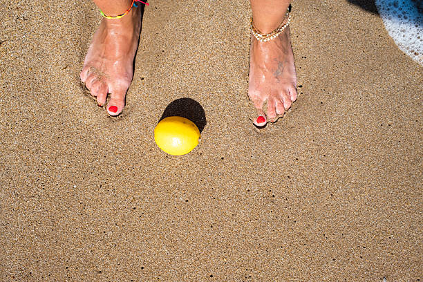 Woman with beautifully painted toenails and stylish anklets standing barefoot on the warm beach, enjoying the sun and relaxing next to a vibrant lemon on a bright summer day