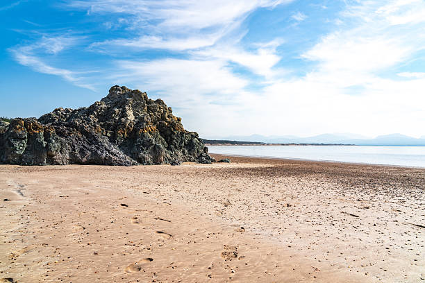Scenic view of footprints on the sandy beach leading to a large rock formation on llanddwyn island, north wales, with mountains in the distance