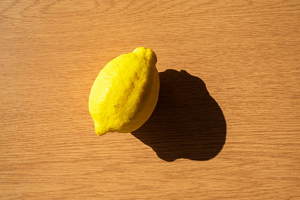 Single, vibrant yellow lemon rests on a light brown wooden table, casting a distinct shadow under the bright sunlight, creating a visually appealing contrast of color and texture