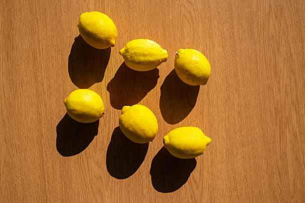 Several ripe yellow lemons are scattered on a wooden table, casting long shadows under the bright sunlight, creating a vibrant and summery scene