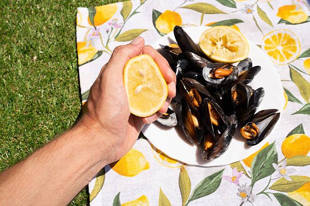 Hand squeezing a lemon half over a plate of succulent cooked mussels, set against a vibrant lemon-patterned tablecloth during a sunny summer picnic on a lush grassy lawn