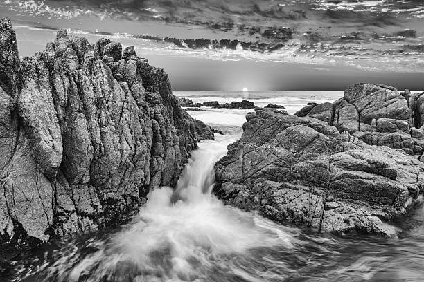 Black and white version of waves rushing between rugged rocks at sunset. Central California coast.