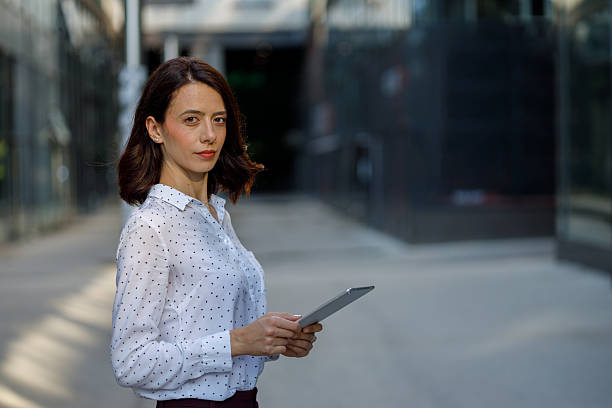 Businesswoman holding a digital tablet stands outside an office building, engaging with technology in an urban environment during the day