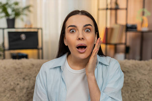 istock Young woman sitting on sofa raising eyebrows opening mouth surprised amazed shocked by unexpected 2229299527