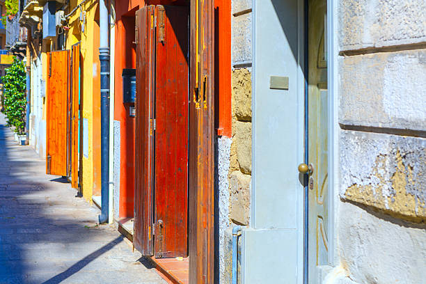 Row of colorful building doors with open wooden shutters lines narrow European street. Bright sunlight creates deep shadows on cobblestone pavement. Folding doors stand out against toned walls