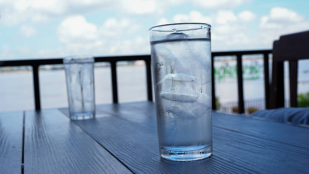 Refreshing glass of water filled with ice sits on wooden table, with another glass in background. scene captures serene outdoor setting by water, evoking sense of relaxation and tranquility