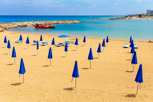 Beautiful sandy beach in Cyprus is lined with many closed blue umbrellas. Calm, turquoise sea is enclosed by a stone jetty. Two small boats are moored near the shore