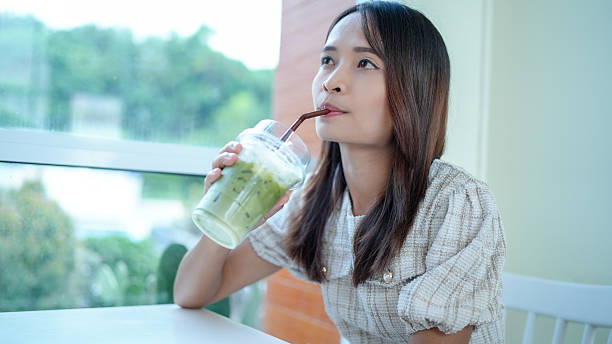 istock Young woman enjoys refreshing green beverage while sitting at table by window, surrounded by serene outdoor view. atmosphere is calm and inviting, perfect for relaxation 2229354082