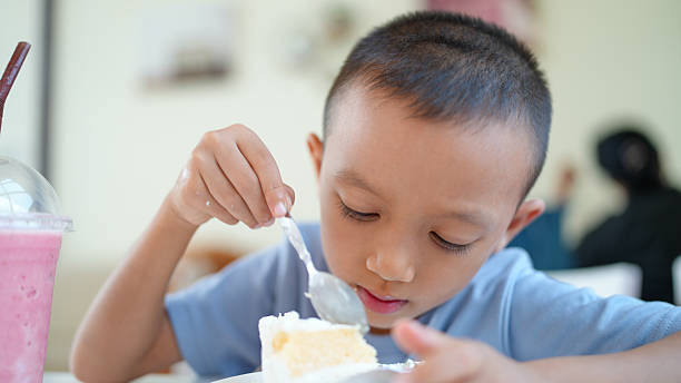 Young boy enjoys slice of cake while sitting at table, focused on his dessert. scene captures moment of joy and indulgence, with refreshing drink nearby