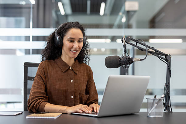 istock A woman smiles while working on a laptop with a microphone setup for a podcast. 2229365558