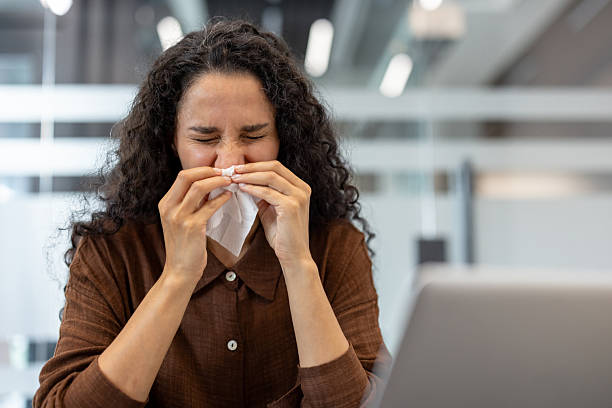 istock A woman with curly hair sneezes into a tissue, likely suffering from a cold at work. 2229365593