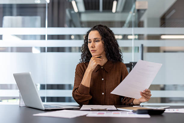 A concentrated businesswoman analyzes paperwork while working on a computer in a modern office setting.