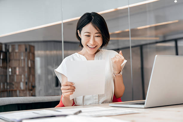 istock Smiling Asian business woman accountant analyst holding documents, doing yes gesture reading good financial report after analysing. Happy eastern businesswoman with paperwork in office. Winner concept 2229394474