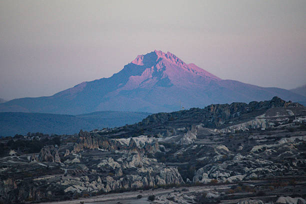 A stunning view of Mount Erciyes glowing in purple light during sunset, overlooking the dramatic rock formations of Cappadocia, Turkey.