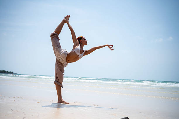 istock Woman with a headscarf performs a standing yoga pose on a tropical beach, balancing gracefully with the ocean in the background 2229409636