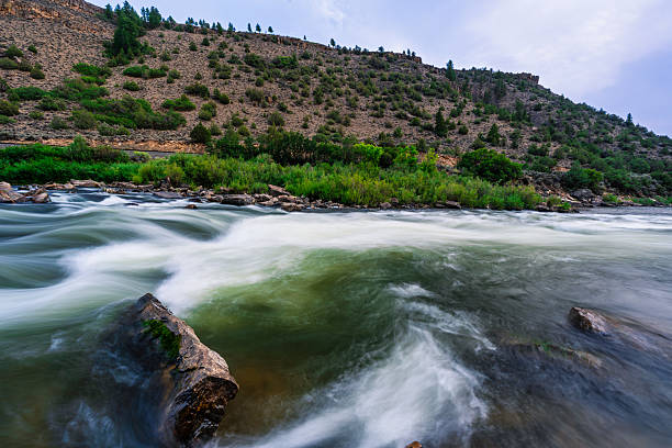 Long exposure photograph of the Colorado River in Colorado, USA. The blurred motion of the rapids contrasts with the sharp, still rocks, highlighting the river’s power and beauty against a backdrop of rugged hillsides and green vegetation.