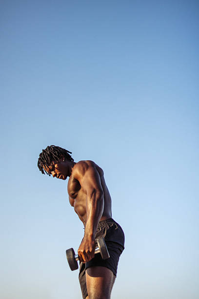 A dedicated young man is performing weightlifting exercises outdoors during a serene sunrise. The tranquil blue sky and natural light enhance the commitment and focus depicted in this moment of physical training.
