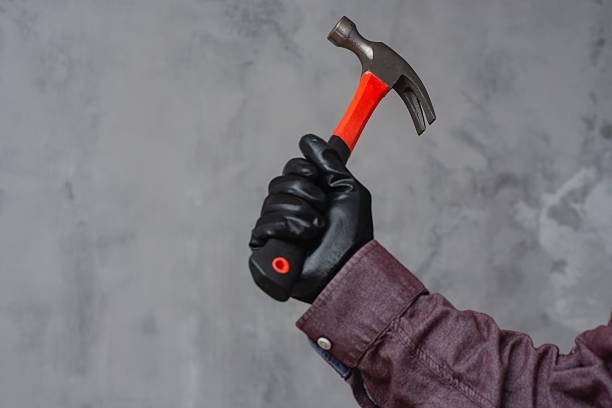 Hand in black protective glove holds hammer with red handle against grey concrete wall. Concept of manual labor, construction works, DIY and repair works