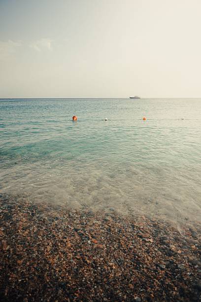 A serene beach scene with pebbles, clear water, and a distant boat under a bright sky.