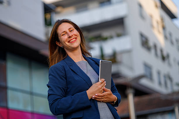 istock Smiling professional woman standing with digital tablet, modern office skyline blending into urban workspace background 2229456228
