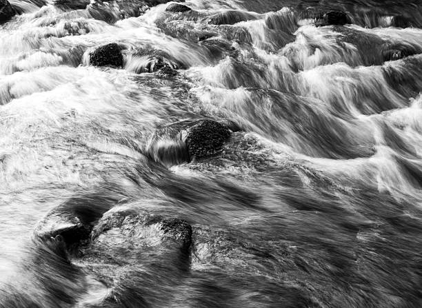 Black and white long exposure of fast-moving river rapids surging around partially submerged rocks, creating smooth patterns and textures in the water