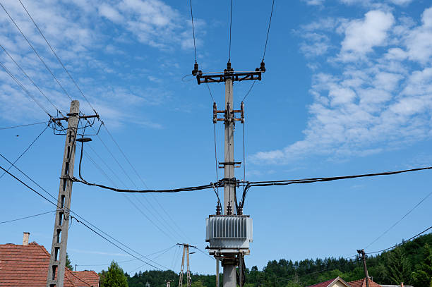 A tall utility pole with an electric transformer and multiple power lines set against a bright blue sky with scattered clouds in a rural setting. The background features rooftops, green trees, and distant forested hills, contrasting man-made infrastructure with nature.