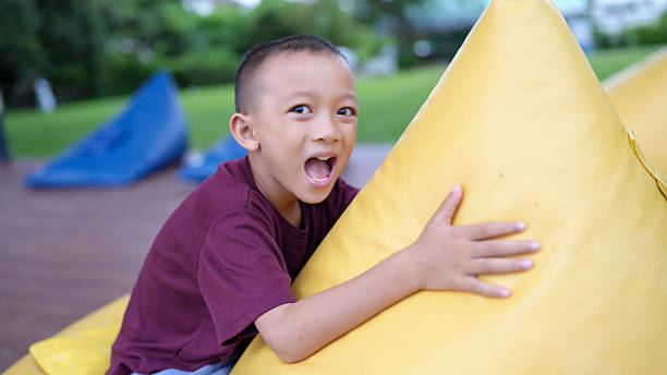 istock Joyful child smiles widely while hugging large yellow cushion, showcasing excitement and playfulness vibrant outdoor setting. background features green grass and colorful bean bags, enhancing 2229501802
